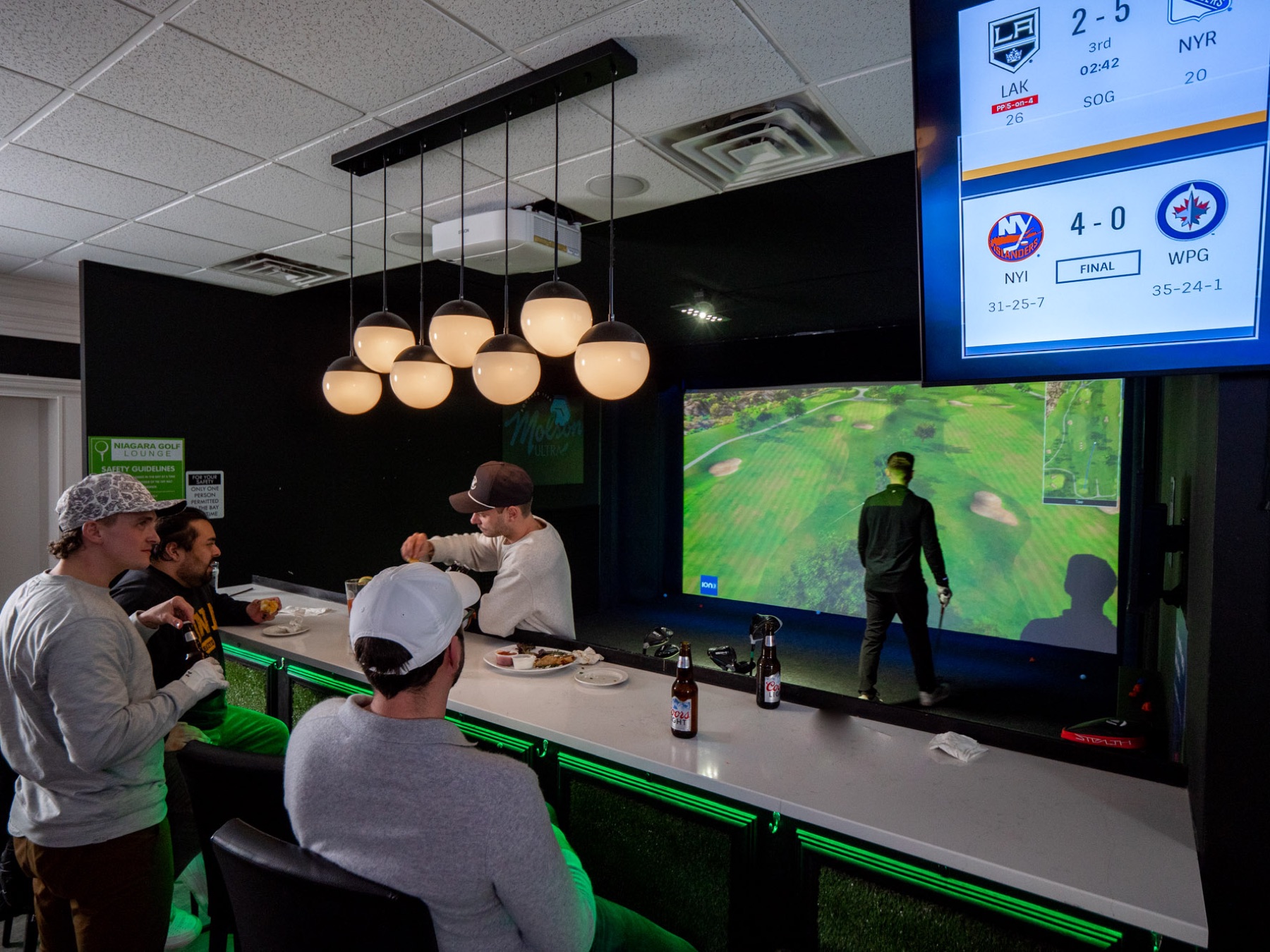 Fans eat and drink along the glowing bay rail while hockey scores play overhead