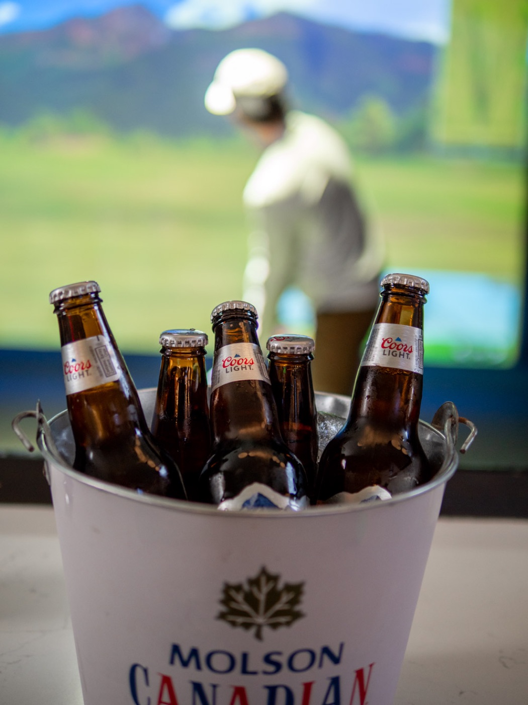 A bucket of cold bottles at the rail while a golfer swings on the simulator behind