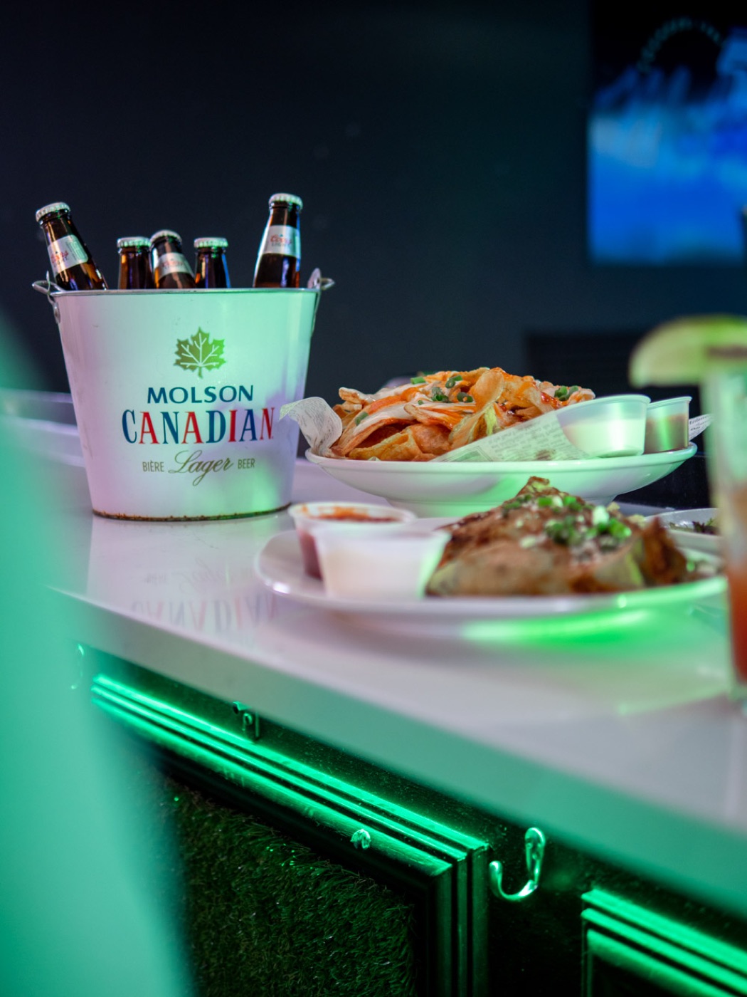 A Molson Canadian bucket and plates glowing on the lime-lit bar