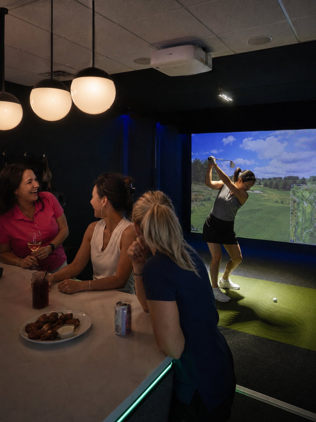 Ladies League night: one player at the top of her backswing in the simulator bay while three friends cheer from the bar