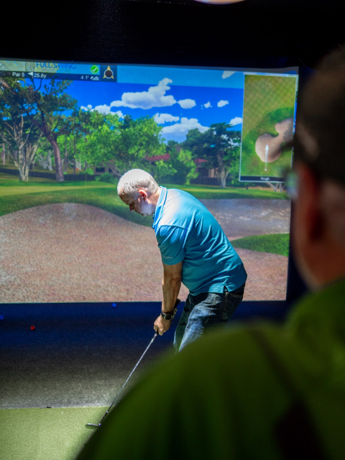 A coach watches over a golfer's shoulder as he lines up a shot in the simulator bay